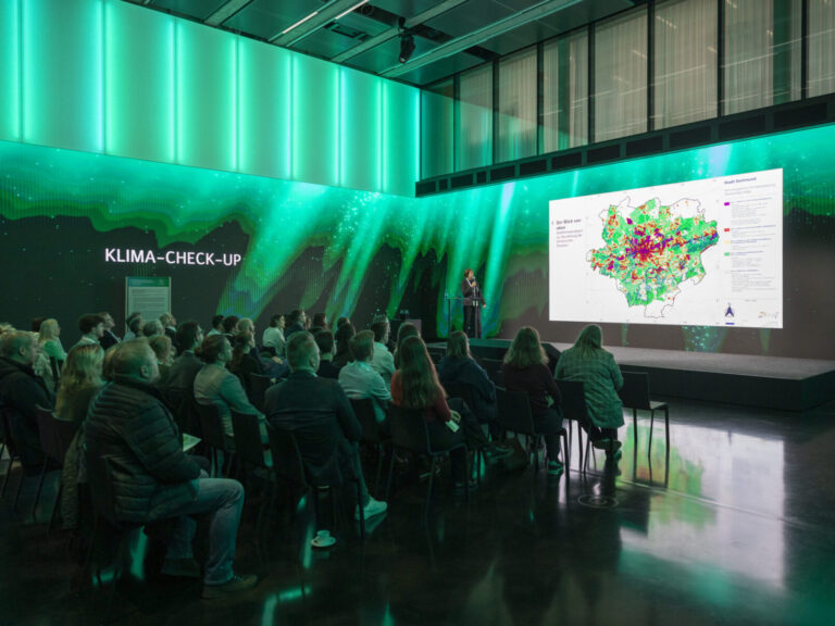 Publikum in grün beleuchtetem Saal guckt in Richtung einer Bühne mit Präsentation einer Karte links steht eine Sprecherin. Links eine Wand mit Aufschrift „KLIMA-CHECK-UP“