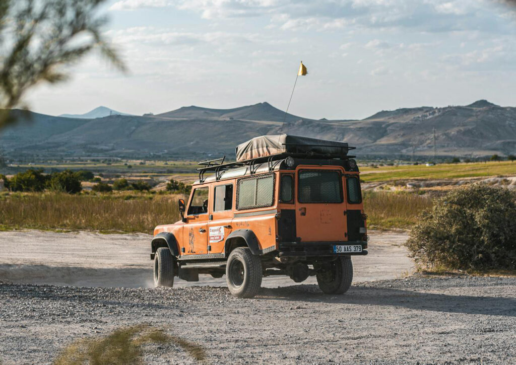 Orangfarbener Geländewagen auf einer Schotterstraße, im Hintergrund eine Feld und Berglandschaft.