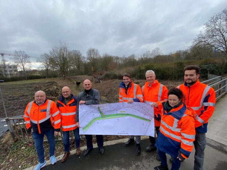 Gruppe von 7 Projektbeteiligten der Emschergenossenschaft, Stadt Dortmund und öffentlich-rechtliche Wasserwirtschaftsverband in neonorangenen Jacken, halten vor Landschaft gemeinsam einen Plan des Emscherverlaufs hoch.