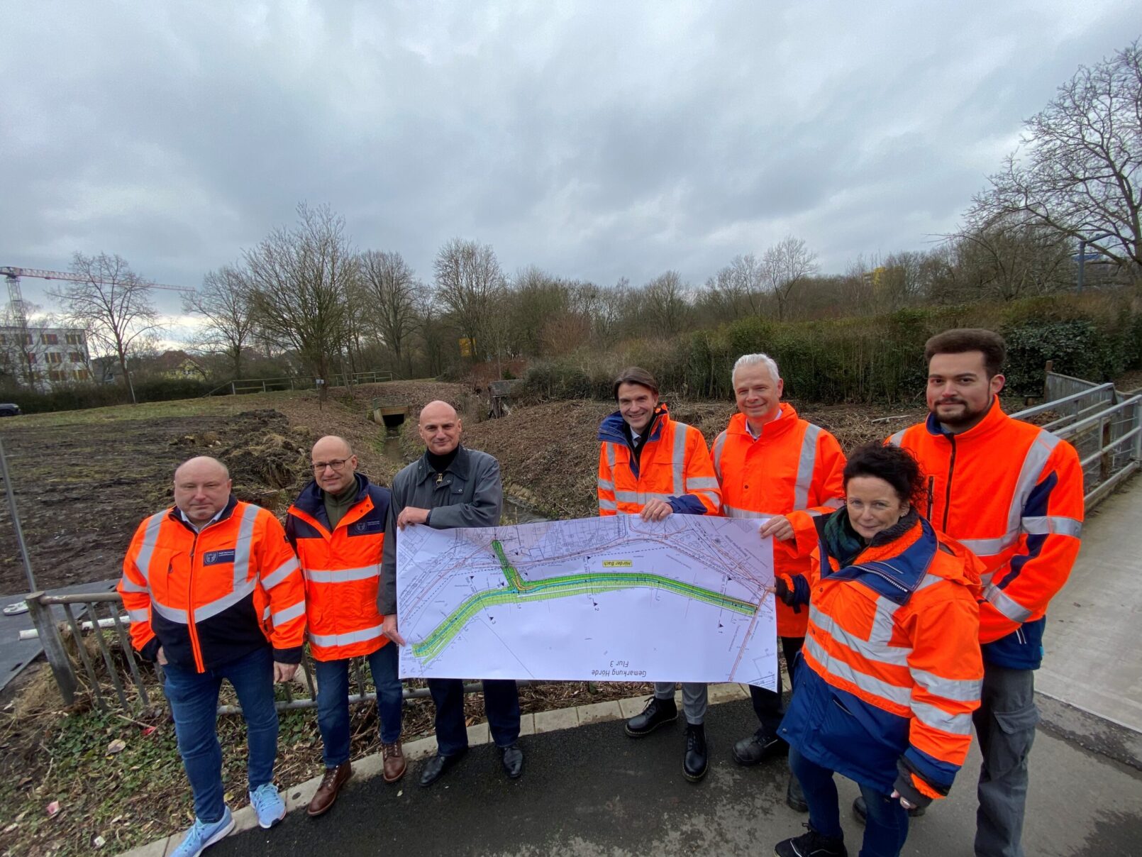 Gruppe von 7 Projektbeteiligten der Emschergenossenschaft, Stadt Dortmund und öffentlich-rechtliche Wasserwirtschaftsverband in neonorangenen Jacken, halten vor Landschaft gemeinsam einen Plan des Emscherverlaufs hoch.