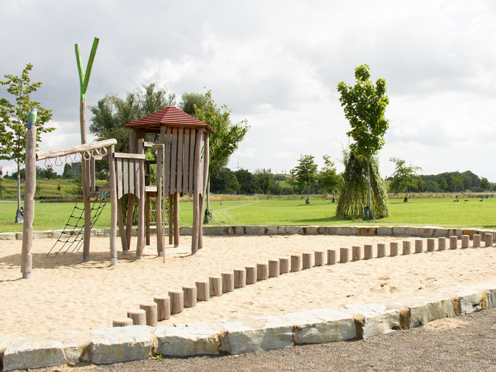 Spielplatz mit Spielgeräten im Vordergrund, Wiese und Weidentipis im Hintergrund des Emscherlandes