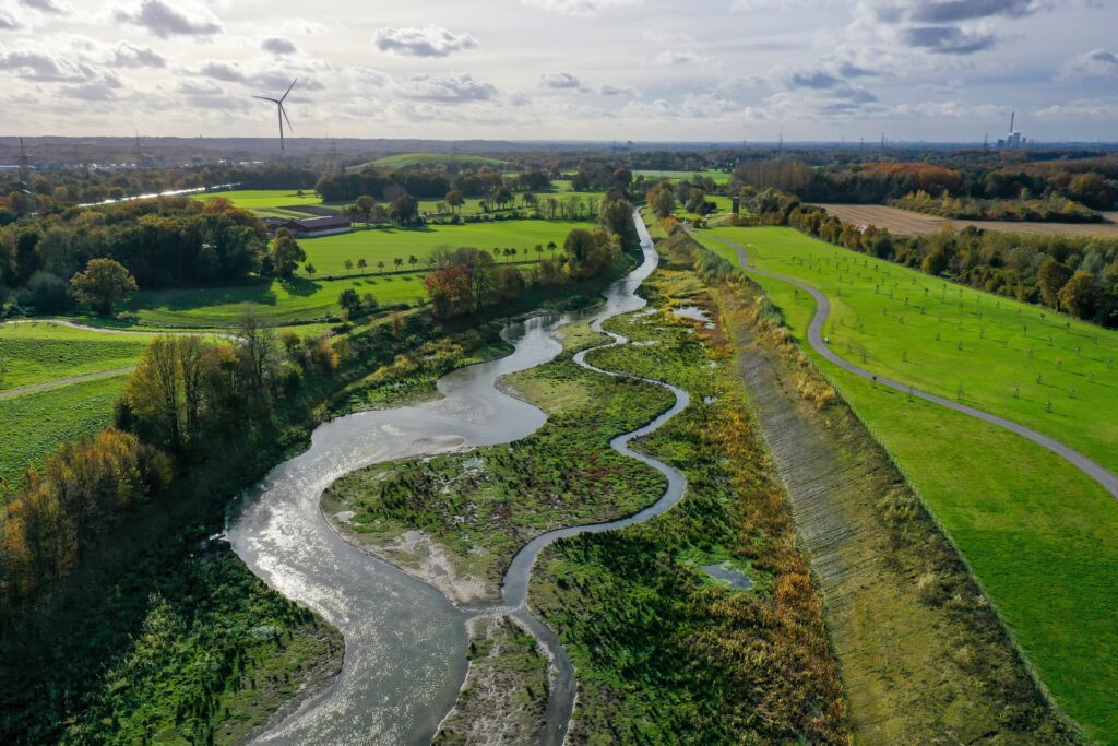 Vogelperspektive auf die Mündung des Suderwicher Baches in die Emscher umgeben von grüner Landschaft. Im Hintergrund das Ruhrgebiet mit Windräsern und Industriegebäuden.